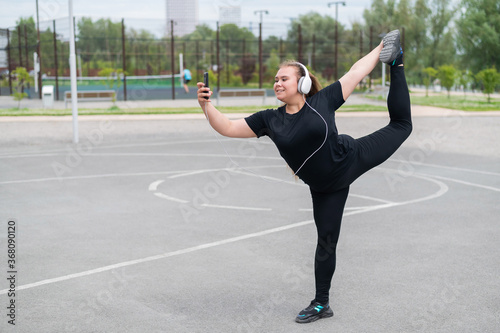 Beautiful fat young woman in headphones makes a figure skater and takes a selfie. A chubby smiling girl listens to music and warms up before jogging outdoors. Sports for weight loss. photo