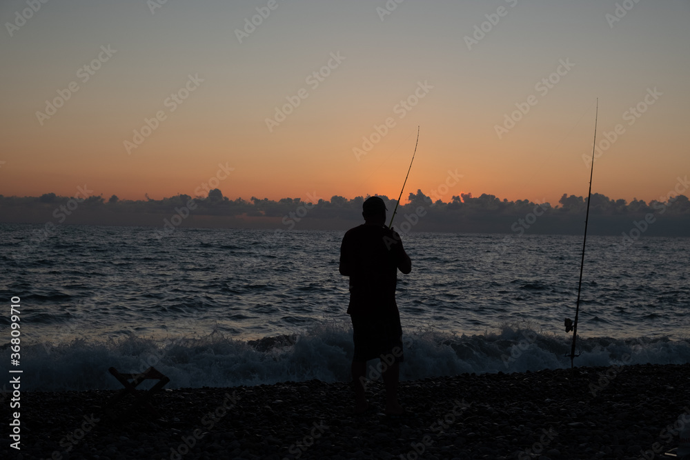 Silhouette of a fisherman against the background of the evening sky. Sunset at the sea. The man is holding a fishing rod. Summer. Georgia. Black Sea.