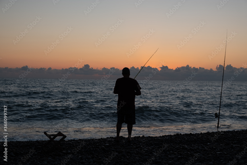Silhouette of a fisherman against the background of the evening sky. Sunset at the sea. The man is holding a fishing rod. Summer. Georgia. Black Sea.