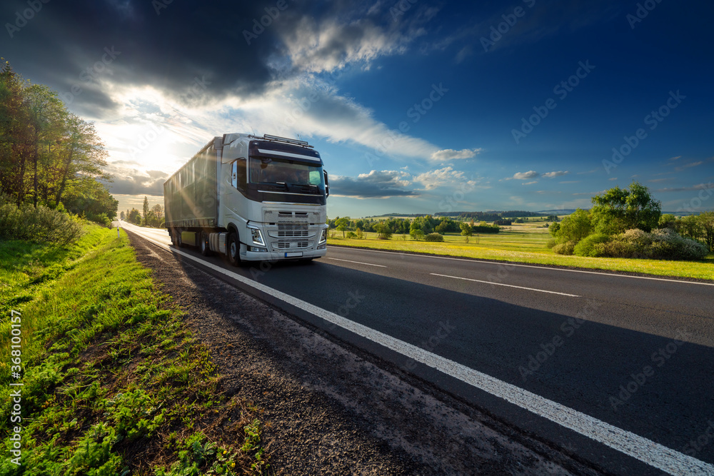 White truck driving on the asphalt road in rural landscape at sunset with dark storm cloud