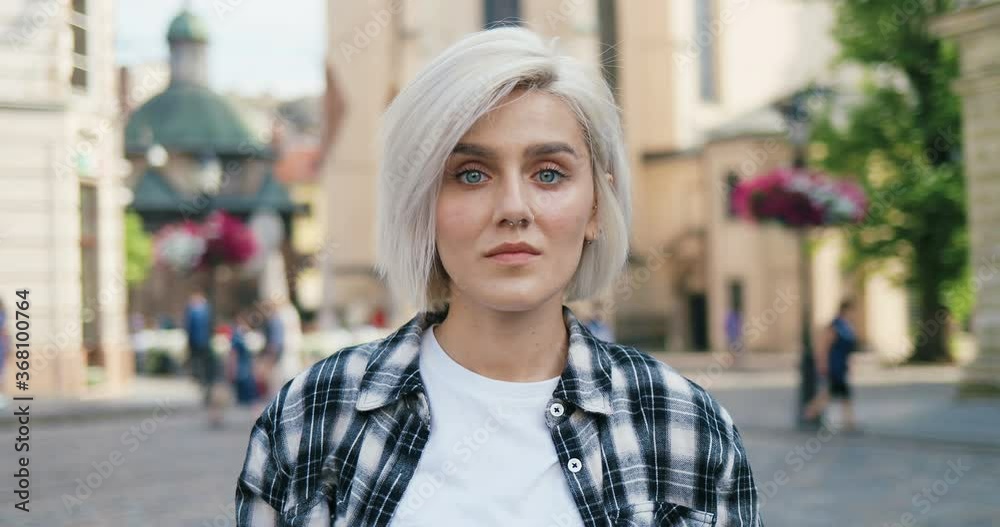 Portrait of young Caucasian blonde girl in plaid shirt and with nose piercing standing in beautiful city outdoor and turning face to camera with smile. Pretty woman in hipster style smiling. Portrait of young Caucasian blonde girl in plaid shirt and with nose piercing standing in beautiful city outdoor and turning face to camera with smile. Pretty woman in hipster style smiling.