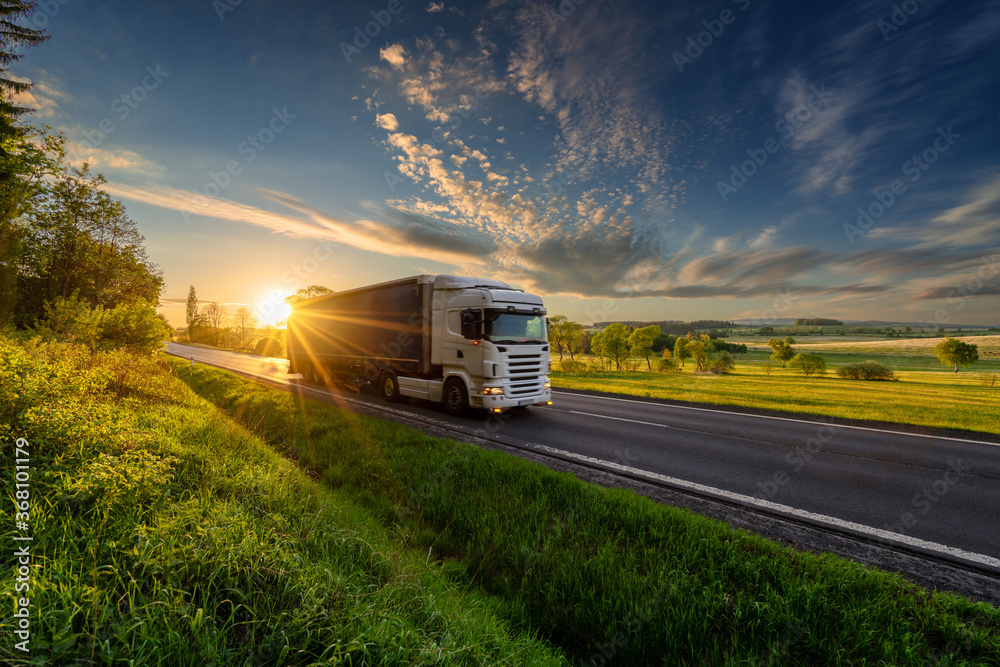 White truck driving on the asphalt road in rural landscape in the rays of the sunset