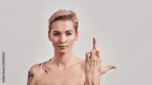 No way. Portrait of arrogant half naked tattooed woman with pierced nose and short hair showing middle finger sign, looking at camera isolated over light background