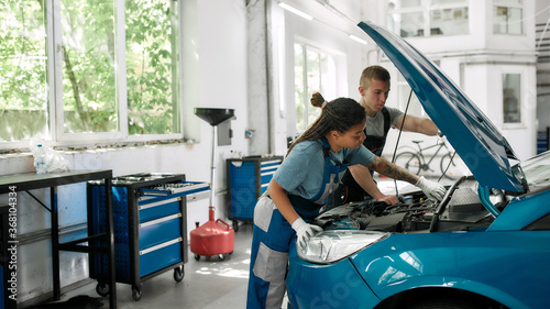 First class performance. African american woman, professional female mechanic repairing car engine, tighten, screw with spanner while her colleague holding torch under car hood at service station