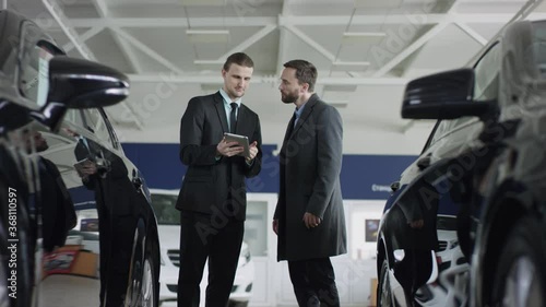 A male bearded client choosing car at dealership with the help of auto seller. Manager in auto dealership presents new cars using a tablet.