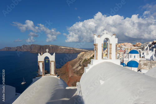 Santorini, Oia - view of the white two bell tower, in the background a church with a blue dome and white houses built on a cliff above the sea.Beautiful blue sky with white clouds.