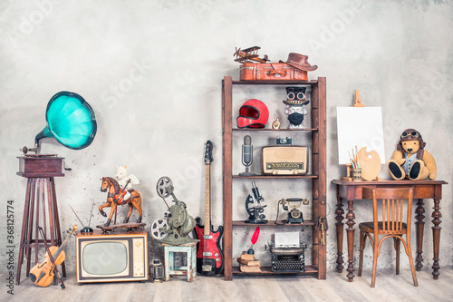 Antique media devices, writers tools, gramophone, film projector, old Teddy Bear toys and white canvas blank on easel, violin and guitar front concrete wall background. Vintage style filtered photo photo