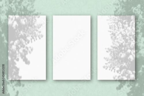 3 vertical sheets of textured white paper on light green table background. Mockup overlay with the plant shadows. Natural light casts shadows from an exotic plant. Horizontal orientation