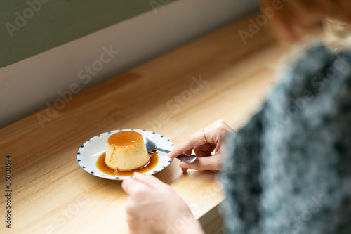 A brown-haired woman wears a green dress, preparing to eat custard pudding, which is placed on a wooden table in a coffee shop.