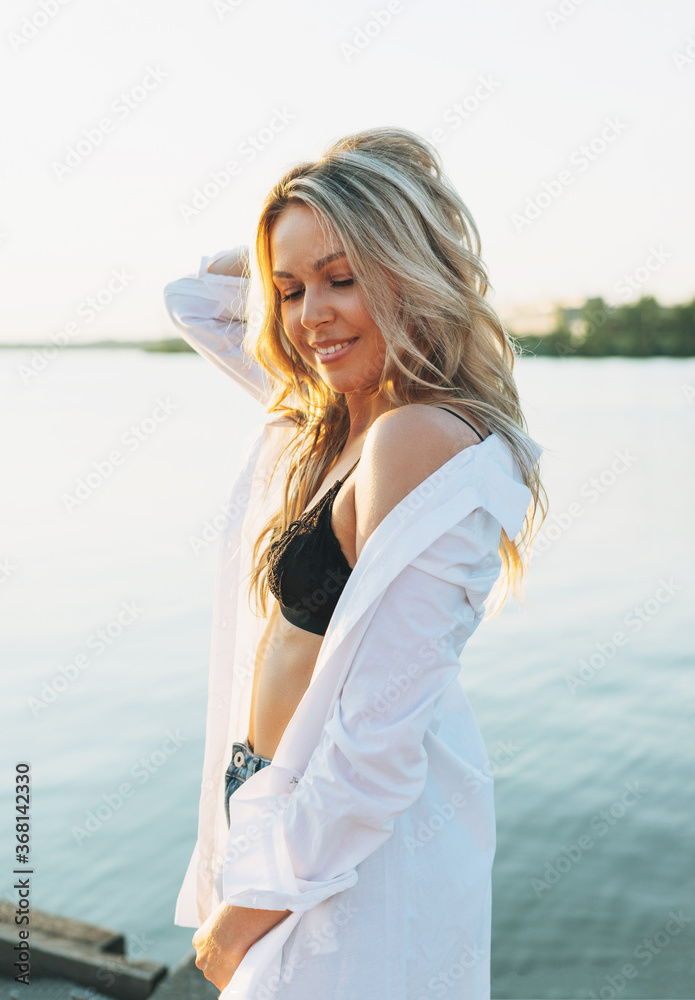 Beautiful smiling blonde young woman in white shirt on the pier on sunset
