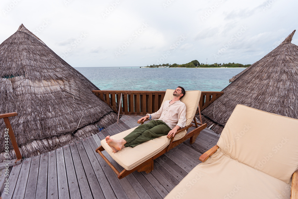 western young man lying on deckchair over water hut at luxury resort in maldives with sea in the background