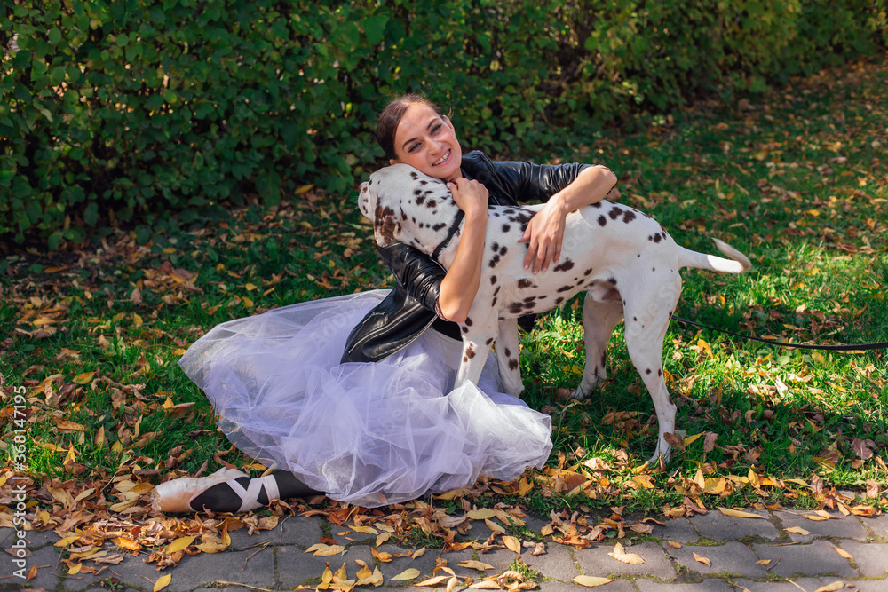Fototapeta premium Ballerina with Dalmatian dog in the Park. Woman ballerina in a white ballet skirt and black leather jacket and in pointe shoes in autumn park hugging her spotty dalmatian dog.