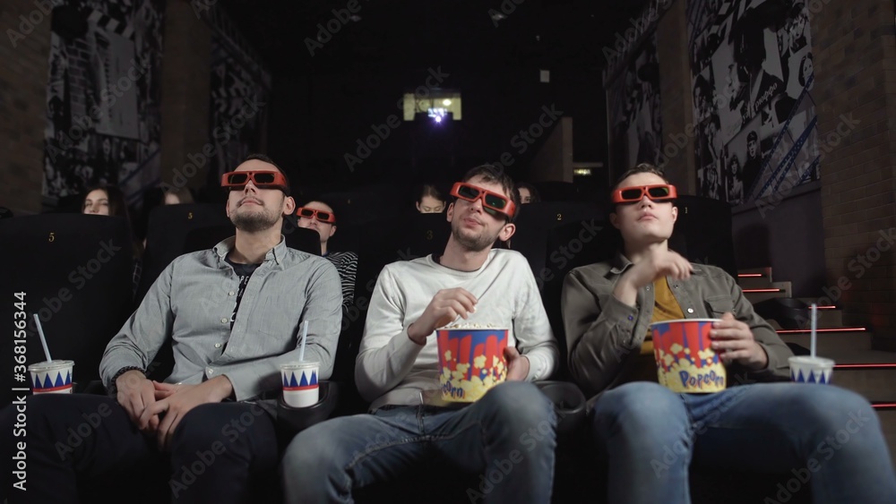 Three friends with 3D glasses are watching a movie and eating popcorn