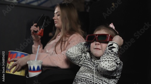 Mother with her daughter are watching 3d film in cinema. Little girl puts on 3D glasses