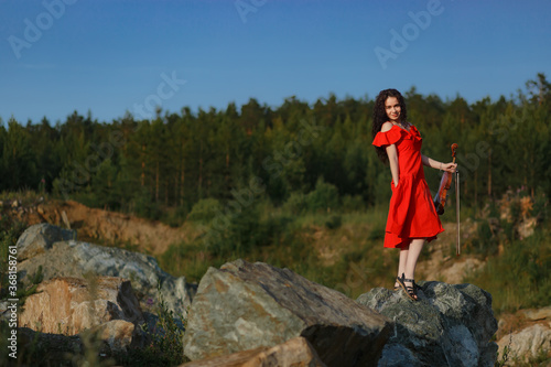 brunette girl with lush curly hair stands on the rocks near the lake in summer in a bright red dress with a violin, professional musician violinist