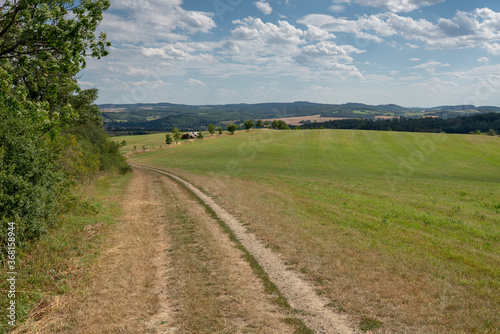 A country road in fields of central Bohemia  with wooded hills on the horizon. There are trees along the road and a pile of logs in the distance. Shot on a summer day  with a partially cloudy sky.