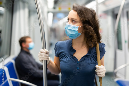 Young brunette in medical face mask and protective gloves riding in modern subway car. Concept of forced city trip in context of coronavirus pandemic..