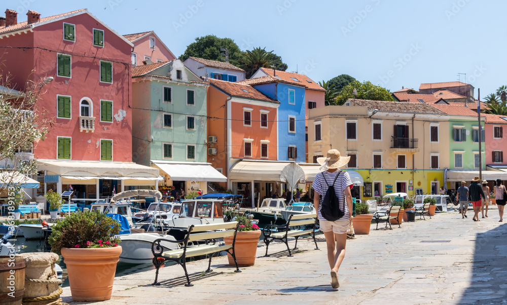Rear view of beautiful blonde young female traveler wearing straw sun hat sightseeing and enjoying summer vacation in an old traditional costal town of Veli Losinj, Adriatic cost, Croatia.