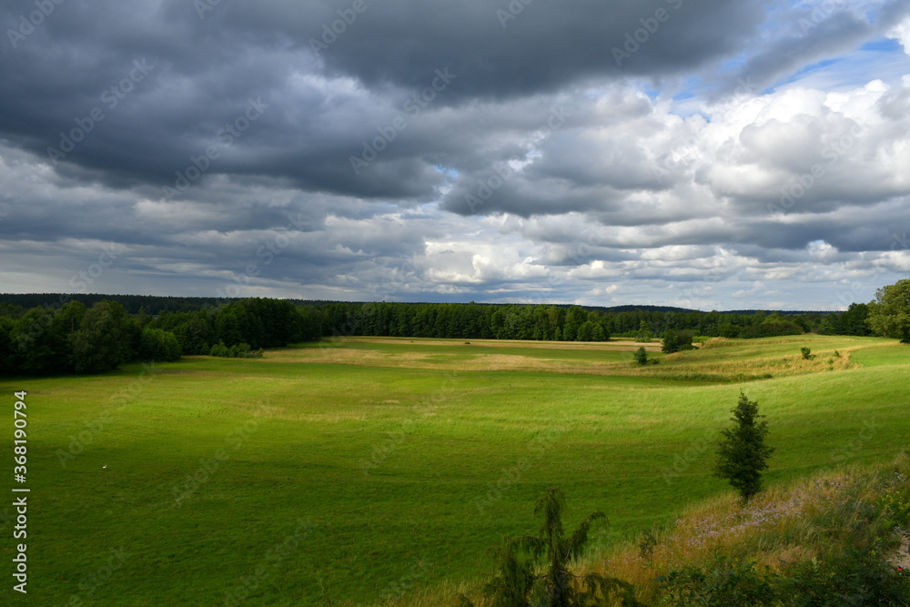 A view of a set of fields, meadows, and pasturelands with some trees and shrubs growing on them seen from the top of a tall hill right before a massive storm with dark, heavy clouds above the horizon 