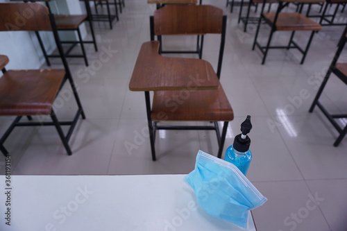 Medical mask and alcohol hand wash gel on the teacher 's desk with wood lecture chairs in the empty classroom. Concept during the Coronavirus Disease COVID-19 outbreak. Back to school concept.