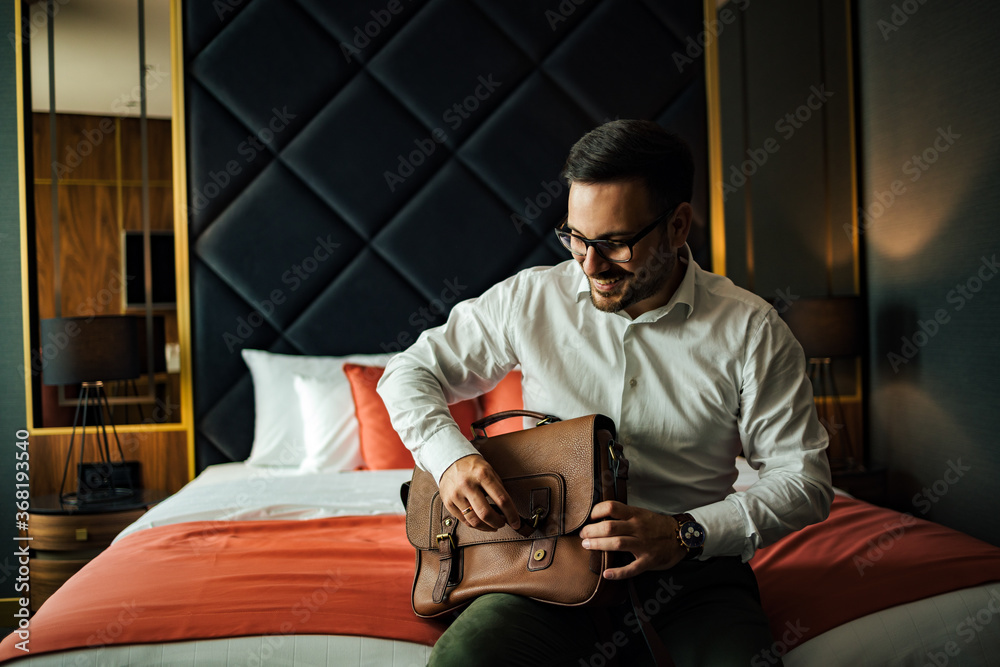 Smiling man with a bag sitting on the bed in the luxurious hotel room, portrait.