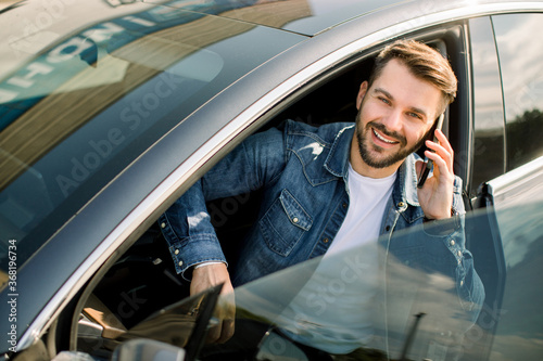 Handsome Caucasian bearded man in casual jeans shirt, sitting in his modern new car, talking on cellphone, smiling and looking at camera. Happy man owner is testing his new car