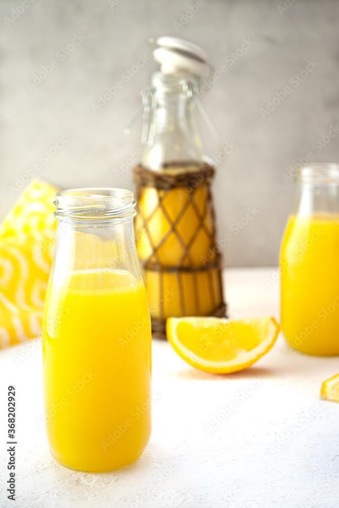 Sweet orange juice in glass decanters and a bottle stands on a white table against the background of a gray concrete wall. One bottle in ethnic style is decorated with rattan. Detox diet, vitamins. Sweet orange juice in glass decanters and a bottle stands on a white table against the background of a gray concrete wall. One bottle in ethnic style is decorated with rattan. Detox diet, vitamins.