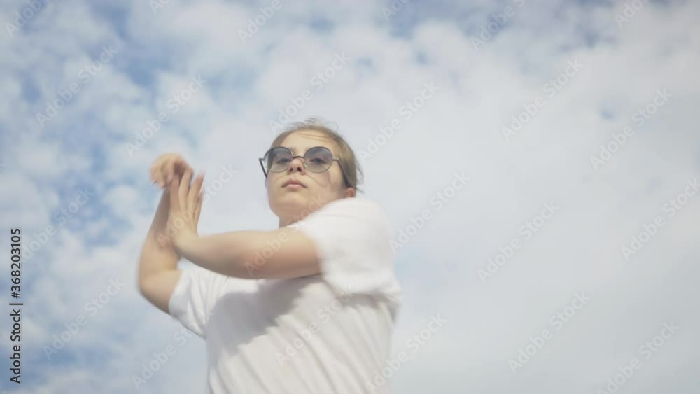 Middle shot of absorbed caucasian girl in eyeglasses dancing at the background of summer sky. Portrait of confident cute Caucasian woman pereforming robot dance moves outdoors. Middle shot of absorbed caucasian girl in eyeglasses dancing at the background of summer sky. Portrait of confident cute Caucasian woman pereforming robot dance moves outdoors.