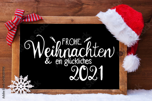 Chalkboard With German Calligraphy Frohe Weihnachten Und Ein Glueckliches 2021 Means Merry Christmas And A Happy 2021. Christmas Decoration Like Santa Hat And Bow. Wooden Background With Snow