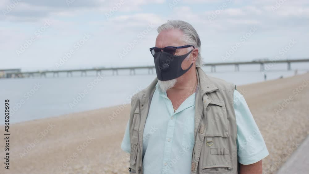 Handheld shot of older man wearing a face mask walking by the sea front, in slow motion