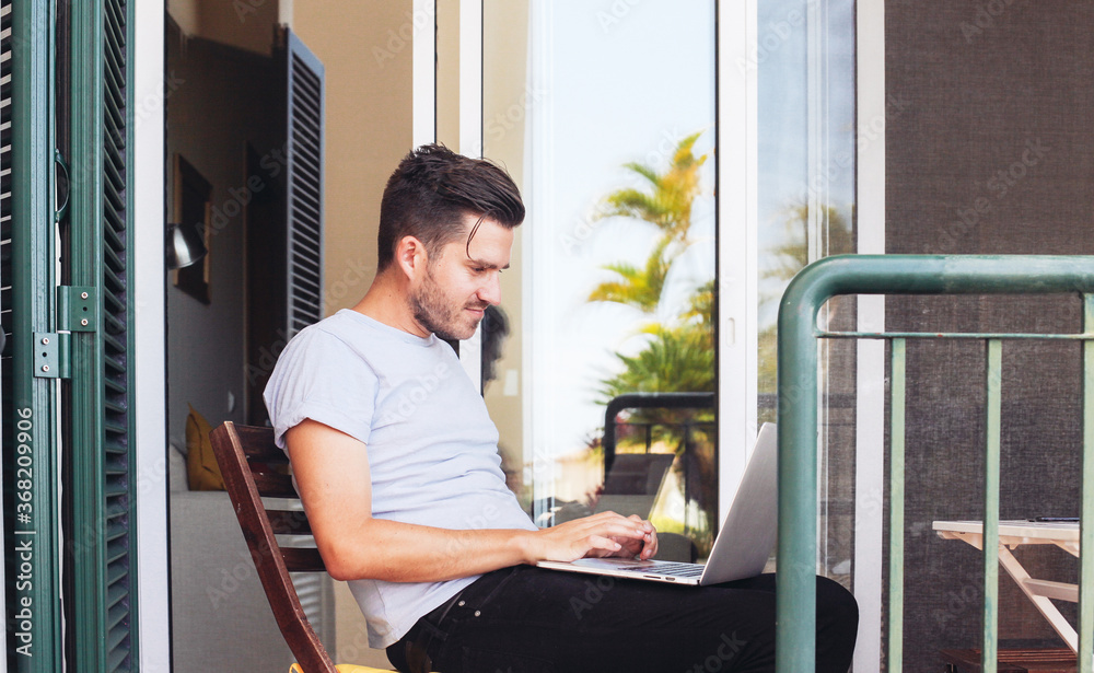 A young man freelancer working on a laptop on a terrace, palms and blue sky on the background. Concept of working remotely