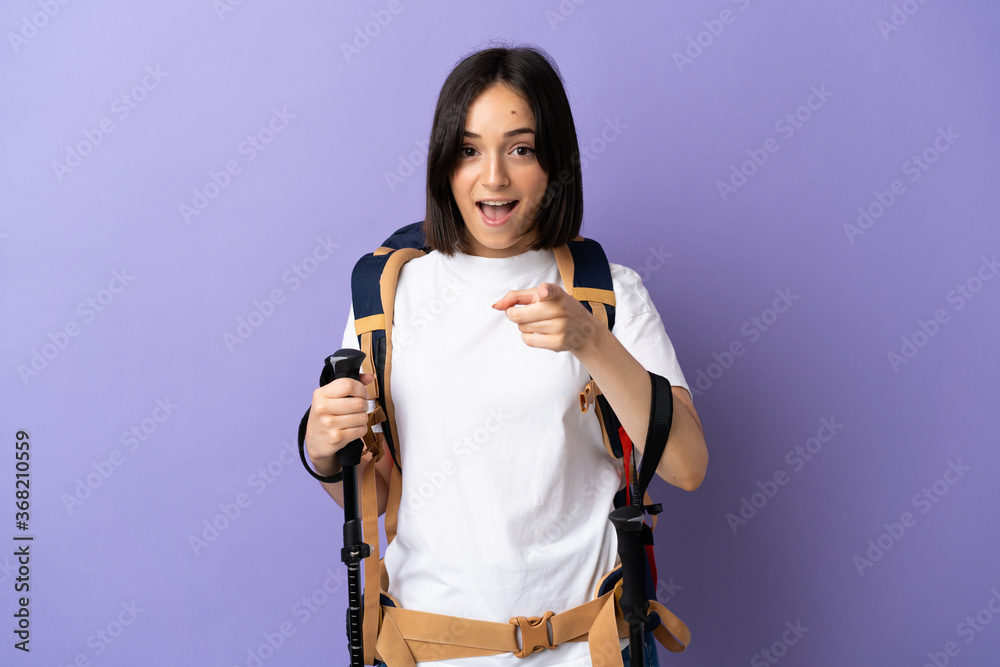Young caucasian woman with backpack and trekking poles isolated on blue background surprised and pointing front
