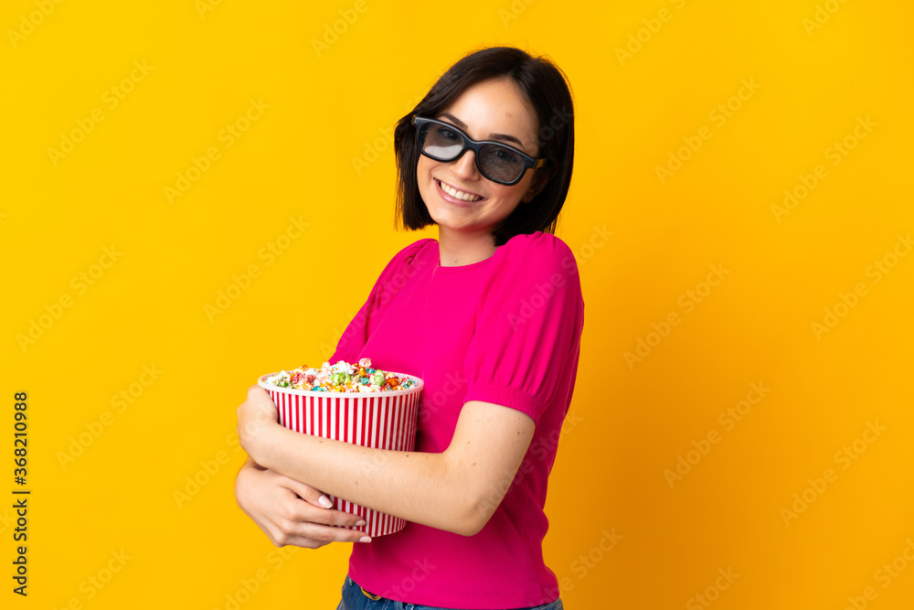 Fototapeta premium Young caucasian woman isolated on yellow background with 3d glasses and holding a big bucket of popcorns