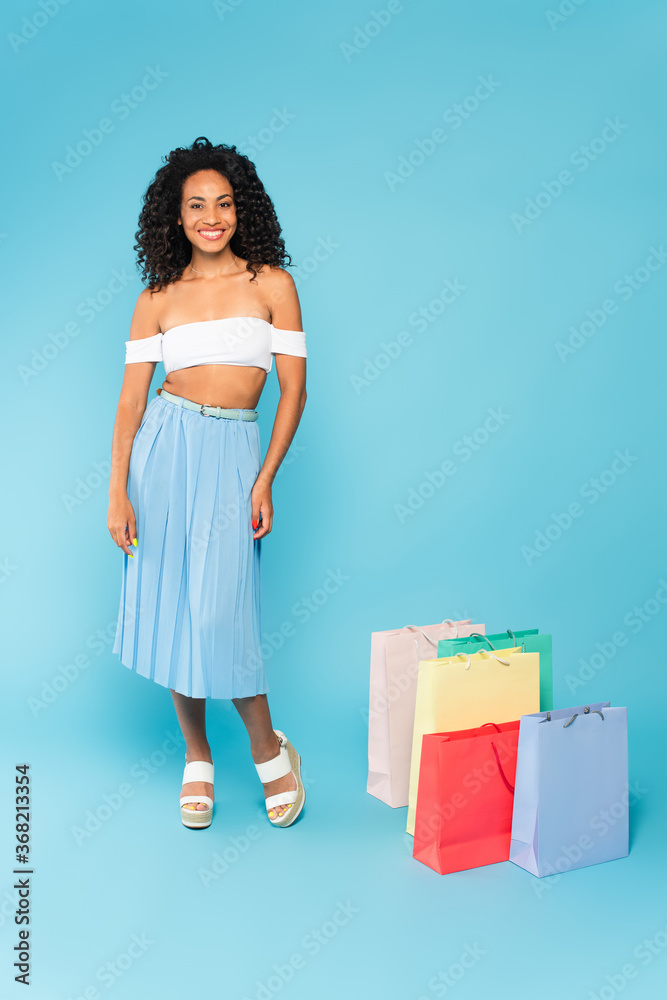 happy african american girl standing near shopping bags on blue happy african american girl standing near shopping bags on blue
