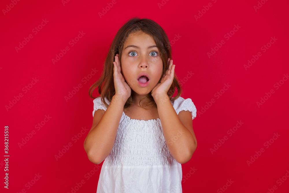 Emotive Little caucasian girl with blue eyes wearing white dress standing over isolated red background with scared expression, keeps hands on head, jaw dropped, has terrific expression. Omg concept Emotive Little caucasian girl with blue eyes wearing white dress standing over isolated red background with scared expression, keeps hands on head, jaw dropped, has terrific expression. Omg concept