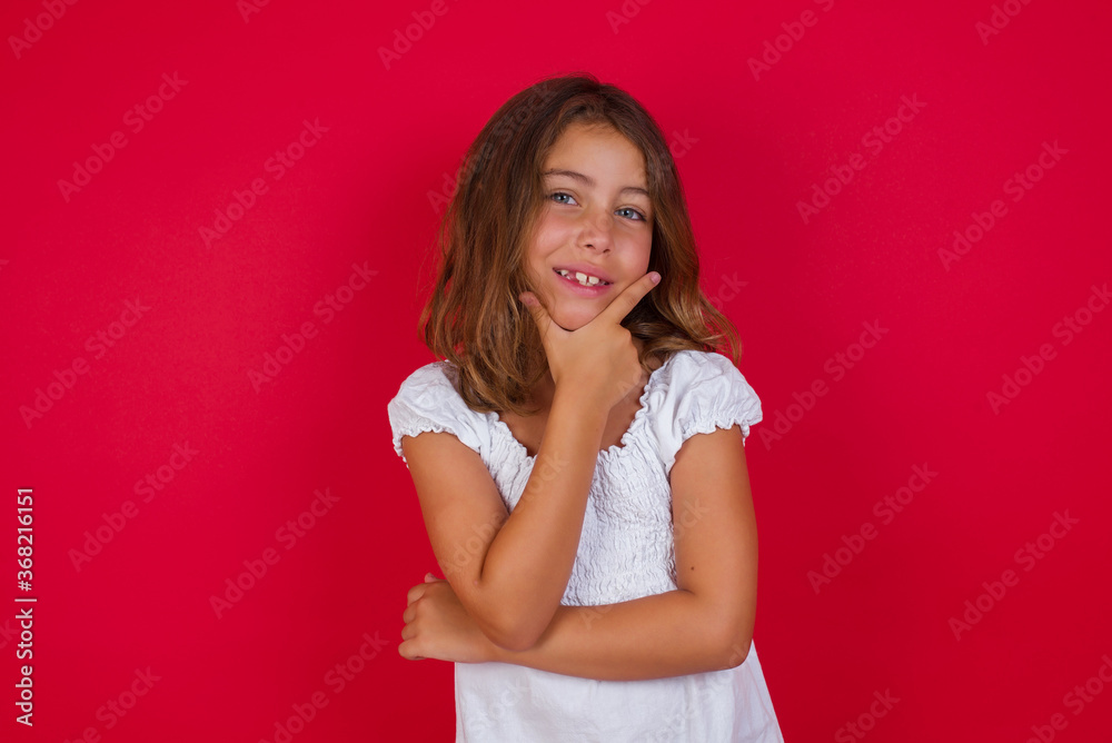 Little caucasian girl with blue eyes wearing white dress standing over isolated red background looking confident at the camera smiling with crossed arms and hand raised on chin. Thinking positive. Little caucasian girl with blue eyes wearing white dress standing over isolated red background looking confident at the camera smiling with crossed arms and hand raised on chin. Thinking positive.
