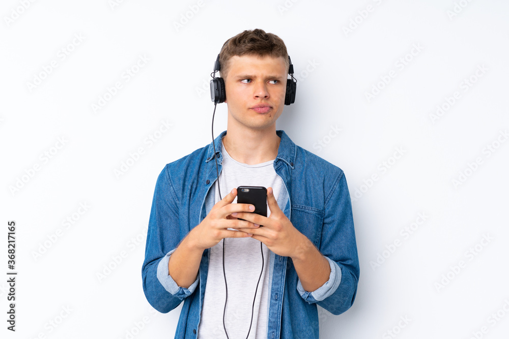 Young handsome man over isolated white background using the mobile with headphones and thinking Young handsome man over isolated white background using the mobile with headphones and thinking