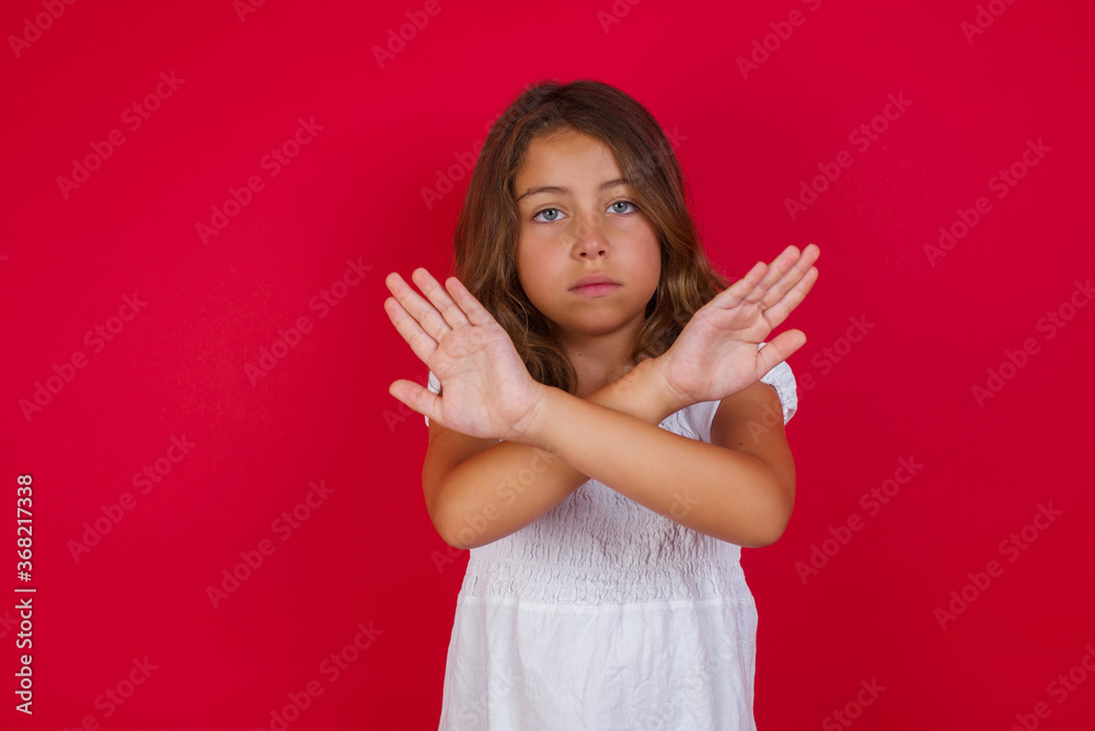 Little caucasian girl with blue eyes wearing white dress standing over isolated red background has rejection expression crossing arms and palms doing negative sign, angry face. Little caucasian girl with blue eyes wearing white dress standing over isolated red background has rejection expression crossing arms and palms doing negative sign, angry face.