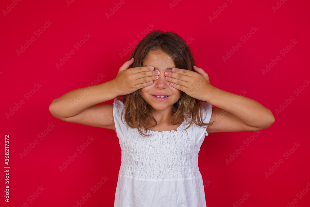 Little caucasian girl with blue eyes wearing white dress standing over isolated red background covering eyes with hands smiling cheerful and funny. Blind concept. Little caucasian girl with blue eyes wearing white dress standing over isolated red background covering eyes with hands smiling cheerful and funny. Blind concept.