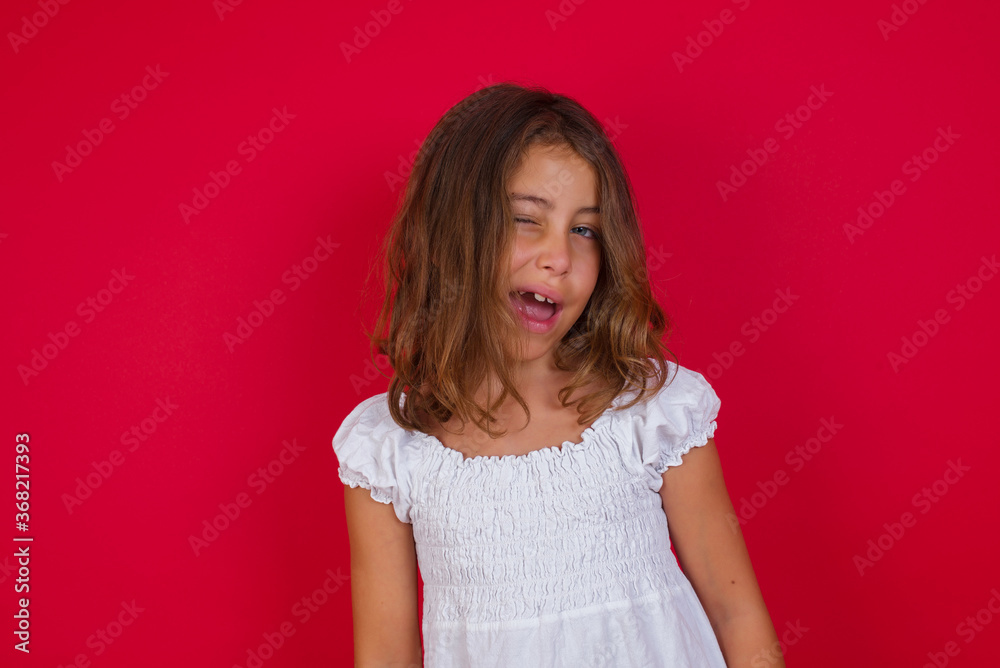 Little caucasian girl with blue eyes wearing white dress standing over isolated red background winking looking at the camera with sexy expression, cheerful and happy face. Little caucasian girl with blue eyes wearing white dress standing over isolated red background winking looking at the camera with sexy expression, cheerful and happy face.
