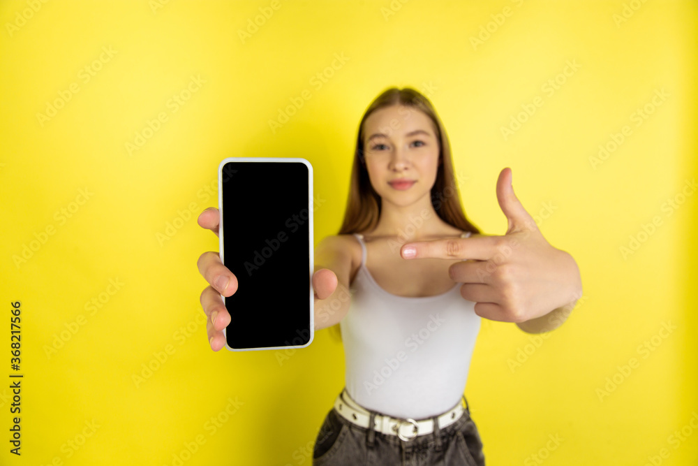 Naklejka premium Pointing on phone's screen. Caucasian young girl's portrait isolated on yellow studio background. Beautiful female blonde model. Concept of human emotions, facial expression, sales, ad, youth culture