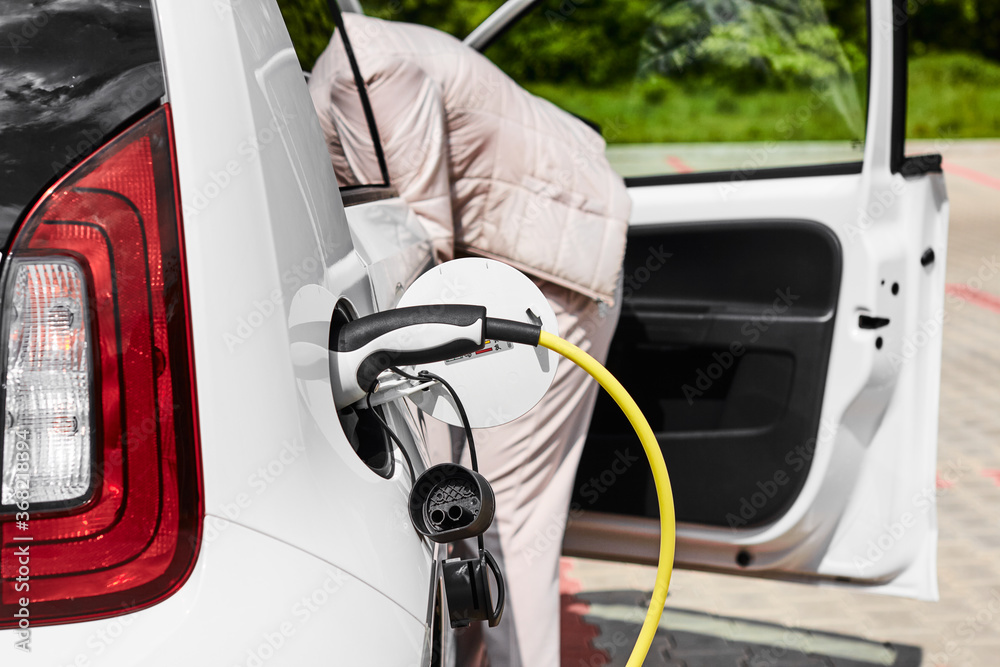 Woman taking something from the car, while electric car is charging at public charging station. Zero emission vehicle Woman taking something from the car, while electric car is charging at public charging station. Zero emission vehicle