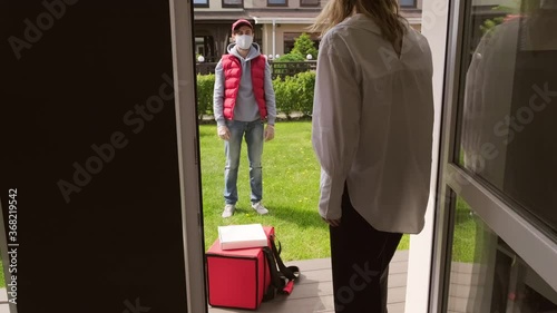 Handheld tracking of blond woman opening her front door and taking pizza box from insulated backpack lying on doorstep as delivery man in face mask waiting photo