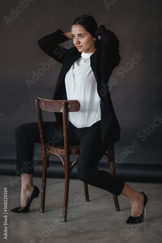 dark-haired girl straightens her hair while sitting on a chair, turned back forward in a black suit, white shirt and black high-heeled shoes