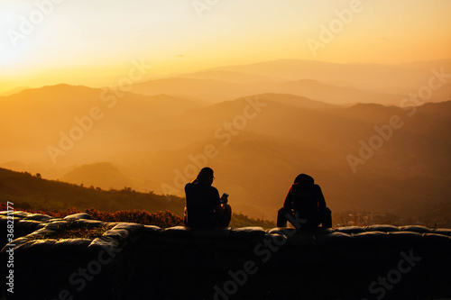 Two tourist young men sitting on rocky cliff and enjoying beautiful sunset view.