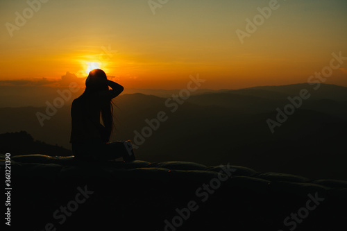 Silhouette of a girl sitting on a cliff side looking at the sunset. Beautiful sunset background.