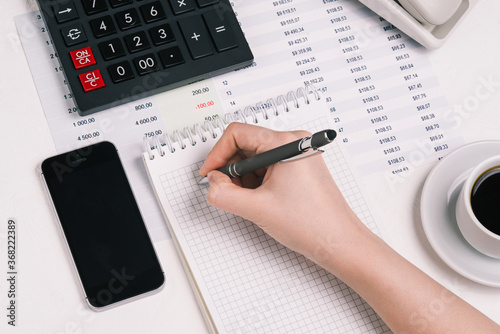 Woman's hand writes in a notebook with a pencil. Desktop with a calculator, mobile phone, coffee Cup and financial documents. Concept of accounting, Finance and Analytics.