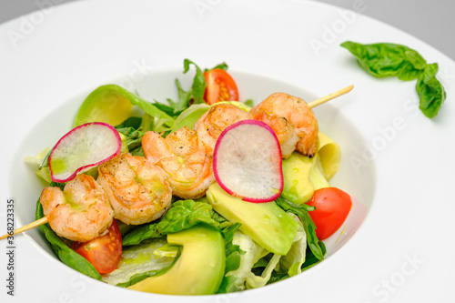Grilled shrimp on a skewer with vegetables on a white plate on a gray background.