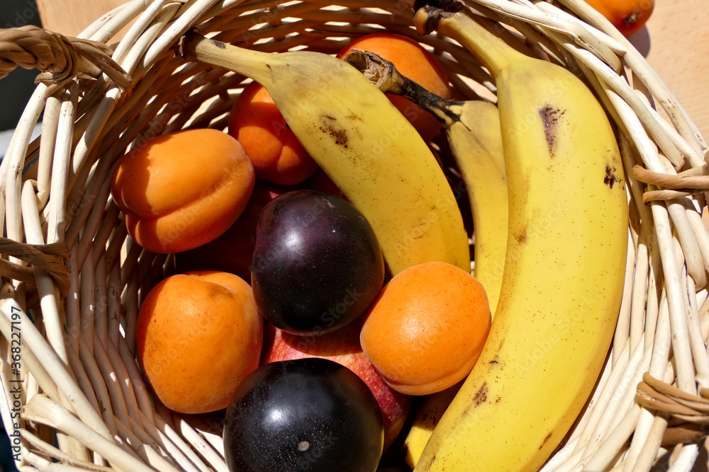 Closeup wooden fruit basket with fresh and healthy bananas, apricots and plums outdoors in beautiful sunlight from above