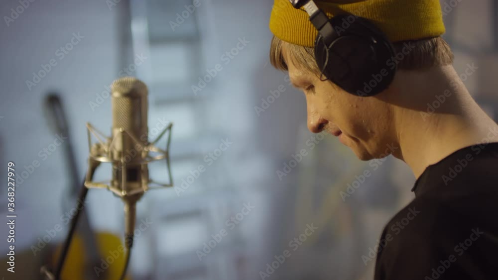 Medium close-up handheld shot footage of young adult man sitting in music recording studio sending messages on smartphone during break